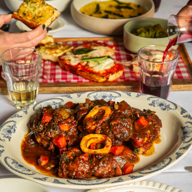 A hearty Italian meal featuring a plate of braised osso buco with vegetables in rich tomato sauce, surrounded by glasses of red and white wine, garlic bread, zucchini, and toasted focaccia with melted cheese and tomato sauce, served on a red gingham tray.