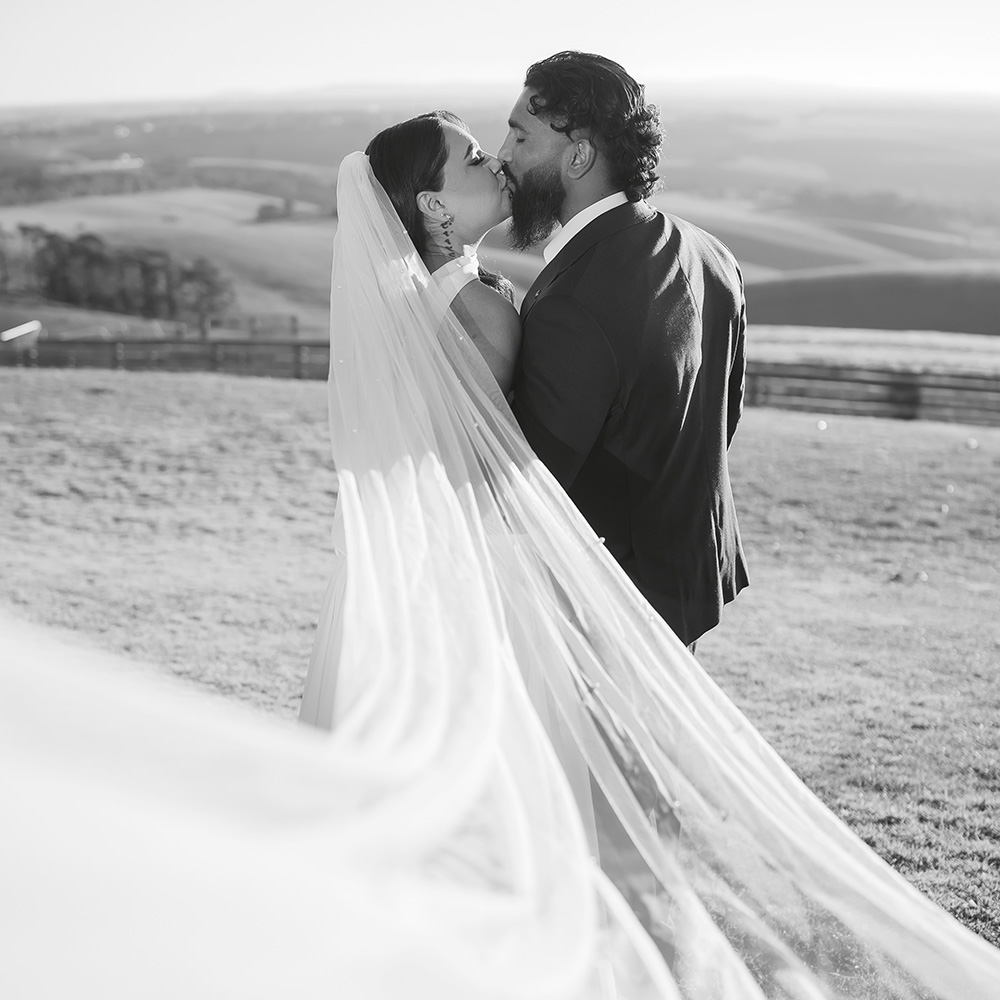 A couple kiss on the hill at Marnong Estate, her veil lifting in the wind, farmland behind them